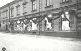 Uppfört av Falkenbergs Sparbank 1895-1896. Indragen butiksentré jämte trappa 1957.(försl. t. bev. pl.)  På bilden finns J E Eliassons AB Manufaktur inrymt i affärslokalen. Övriga affärer är Rika Skoog Hatt- & Möss-Affär, Alma Olsson Frukt Konfektyr.