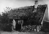 Kvinna framför sin torparstuga med vasstak