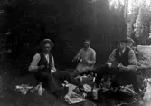 Tre män har lunchrast i skogen. Termotsar, mjölkflaskor och smörgåsar är uppdukade på marken.