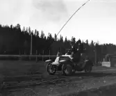 I full fart kör bilen fram på en grusväg. Mannen sitter vid ratten och kvinnan bredvid och håller hårt i sin hatt.
