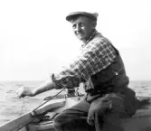 En man i keps, rutig skjorta, väst och byxor sitter i en eka till sjös och dörjar.