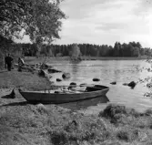 Rogbergasjön. En eka ligger uppdragen vid strandkanten. I bakgrunden sitter en baklädd man på en sten och samtalar med två andra män.