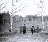 Foto av två män stående framför en grind som omges av två björkar hållandes någon form av mätverktyg i händerna. Text i album: 