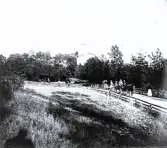 Foto av en grupp människor som går och åker häst med vagn på en väg, i bakgrunden bakom träden syns en byggnad av något slag, någonstans i Sverige