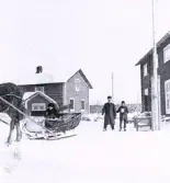 Foto av en häst med släde där det sitter en person, två militärer på skidor till höger i bild samt byggnader i bakgrunden i ett vintrigt landskap. Text i album: 