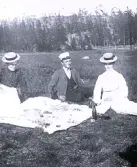 Foto av tre personer som har picknick på ute i det gröna. I bakgrunden syns skog och en gärdsgård.