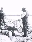 Foto av två män som står vid en stenig strand eller klippor vid vattnet.