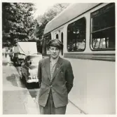 En busschaufför i rutig skjorta, kavaj och skärmmössa står utanför sin buss, trol. någonstans i närheten av
järnvägsstationen, Växjö.