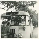 En busschaufför i kavaj och skärmmössa står framför sin buss, trol. någonstans i Växjö.