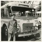 En chaufför i rutig skjorta, kavaj och skärmmössa står vid sidan av en SJ-buss, trol. i närheten av järnvägsstationen, Växjö.
På skylten kan man läsa: 