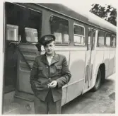 En chaufför i kavaj och skärmmössa, med cigarett i handen, står vid sidan av en SJ-buss, trol. i närheten av järnvägsstationen, Växjö.