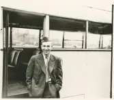 En glad chaufför i kavaj, slips och båtmössa står vid sidan av en buss, trol. i närheten av järnvägsstationen, Växjö.