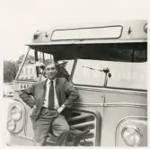 En chaufför i kavaj och båtmössa poserar framför en buss, trol. i närheten av järnvägsstationen, Växjö.