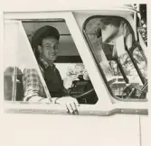 En glad chaufför i rutig skjorta, väst och skärmmössa tittar ut från en buss, trol. i närheten av järnvägsstationen, Växjö.