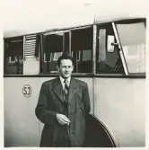 En chaufför klädd i rock med kostym och slips står invid en SJ-buss med en cigarett i handen, trol. på busstorget/järnvägsplan vid järnvägsstationen, Växjö.