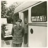 En chaufför i uniformskavaj (med sorgband) och skärmmössa står vid sidan av en SJ-buss på Norra Järnvägsgatan, i närheten av busstorget/järnvägsplan i Växjö. I bakgrunden skymtar gångbron över järnvägen.