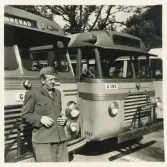 En chaufför i kavaj och båtmössa poserar ledigt framför några SJ-bussar, trol. på busstorget/järnvägsplan i Växjö.