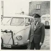 En medelålders man i kavaj, väst och skärmmössa står bredvid en Volkswagen-buss. I bakgrunden skymtar några andra bussar och
en tegelbyggnad, trol. i närheten av järnvägsstationen, Växjö.