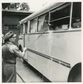 En man i väst och skärmmössa är sysselsatt med att tvätta fönstren m.m. på en SJ-buss.
I bakgrunden skymtar ett bussgarage.