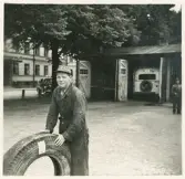 En yngre man i overall och keps, med ett däck framför sig, har stannat upp framför bussgaraget, Växjö.
I bakgrunden syns entrén till dåv. hotell Savoy på Norra Järnvägsgatan.