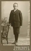 En okänd pojke i mörk kostym med kortbyxor poserar vid en s.k. bankett hos fotografen.
I nedre högra hörnet syns ett inpräglat årtal: 