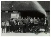 Tröskning i Ågård, Tvååker. Tröskning med fotogenmotor s k tändkulemotor, 1910. Tröskfolket, med barn och vuxna, är samlat omkring maskineriet. På väggen till ladan bakom folket sitter ett stort anslag. 

Från vänster: 1 Okänd. 2 Svante Svensson, son till Sven Bengtsson Ågård. 3 Sven August Bengts-son, ägare till gården. 4 Karl Svensson, son till Sven August Bengtsson. 5 Birger Johannesson, son till Sofia Svensson Ågård. 6 Olga Svensson, dotter på gården. 7 Sofia Svensson  Ågård. 8 Jenny Svensson, dotter på gården. 9 Dräng Birger Karlsson (Eriks Birger) senare bosatt i Tyllered. 10 Sven Johannesson, son till Sofia Svensson. 11 Arvid Johansson, Källdalen, Tvååker. 12 Lars Andersson (Lillebonna-Lars) maskinist. 13 Oskar Johansson, gift med Maries Sofia i Björkäng. 14 Sven Andersson, ägare till gård i Smurte. 15 Johan Andersson, Rönnås, senare ägare till 