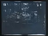 Ett stort sällskap fikar vid ett bord, utomhus bland tallar. Fotografens anteckning på negativet 