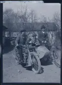 Tre rökande män på en gårdsplan. En på motocykel med en vän i sidovagnen, den tredje på cykel.