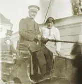 En kusk i livré tillsammans med en ung kvinna, har stannat upp sitt ekipage (en charabang) för fotografen. 
Bakom dem sitter en man i skärmmössa och kavaj.