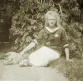 En tonårsflicka i sjömansblus sitter i en trädgård tillsammans med en sovande hund.