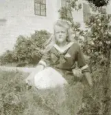 En tonårsflicka i sjömansblus poserar i en trädgård utanför ett större, slottsliknande hus (Skoklosters slott?).