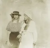 Två tonårsflickor poserar för fotografen utomhus. Den ena i randig blus, stråhatt och mörk kjol, den andra i ljus sjömansklänning och spetshätta.