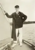 En ung man i seglarkostym med kritade vita skor, poserar i fören på en segelbåt.