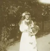 En glad tonårsflicka i ljus sjömansklänning poserar för fotografen en solig sommardag. I famnen ser hon ut att ha några baddräkter (?).