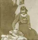 En glad tonårsflicka i blus och kjol sitter på en vedhög på en solig bakgård.