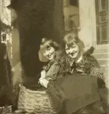 Två tonårsflickor (systrar ?) i blus och kjol vid en vedkorg på en bakgård.