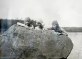 Tre tonårsflickor i baddräkt, till hälften gömda bakom ett stenblock vid en sjö.