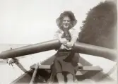 En ung kvinna i randig blus med sjömanskrage, mörk kjol och stråhatt är ute och ror en eka på en sjö, en
varm sommardag.