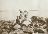 Fem tonårsflickor i ljusa sommarkläder och en pojke har slagit sig ner på några stenar vid en sjökant.
Helgasjön (?).