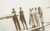 Fyra tonårsflickor i ljusa kläder och två tonårspojkar (varav den ene tänder en cigarett) på en brygga vid Helgasjön (?).