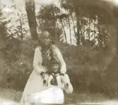 Två unga kvinnor i vita sommarkläder med sjömanskrage, i utkanten av en trädgård.