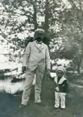 Stadsfiskal Filip Rehnström i sommarkostym med en av hans pojkar i 2:a äktenskapet, trol. Nils Gustaf R. (1903-1993) eller Sven Göte R. (1905-1949), vid en sjökant trol. på Evedal, Växjö. I bakgrunden syns en del av en brygga med några båtar.