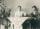 Fru Elisabeth Bergstedt, dottern Margareta och fru Mathilda Rehnström sitter på en dyschatell med ett spelbord framför sig.
På bordet syns ett syskrin med sammetsklädsel.