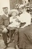 Kronolänsman Albert Bergstedt med dottern Margareta och frun Elisabeth sitter på en bänk i trädgården utanför ett bostadshus.