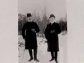 Snöbild. Två herrar i kubb tar varsin cigarett under en promenad i vinterlandskap. Fotograf okänd.