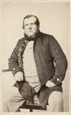 En medelålders man i länsmansuniform, hos fotografen. Han sitter på en stol och lutar ena armen på ett pelarbord.
Kronolänsman Johan Magnus Wilhelm Eksandh, Nottebäck, senare Slätthög.
Mormors far till givaren.