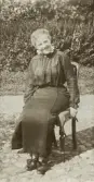En leende kvinna i mörk blus och kjol sitter på en stol i solskenet på en gårdsplan.
Elisabeth Eksandh (senare g. Bergstedt), Slätthög. Mormor t. givaren.