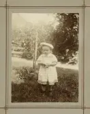 En liten flicka i tvärrandig klänning med invävt mönster och ljus sjömansmössa poserar för fotografen i en trädgård.
Margareta Bergstedt (senare g. Lidén), Växjö.