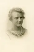 En ung kvinna i ljus klänning med s.k. båtringning. Runt halsen syns en halskedja. 
Präglat fotografnamn m.m. i nhh. 
