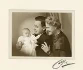 En man och en kvinna med en baby i dopdräkt.
Midjebild. Ateljéfoto.
Oläslig fotografsignatur i nedre högra hörnet.

Carl Rudolf och Margareta Lidén med dottern Elisabeth på dopdagen.